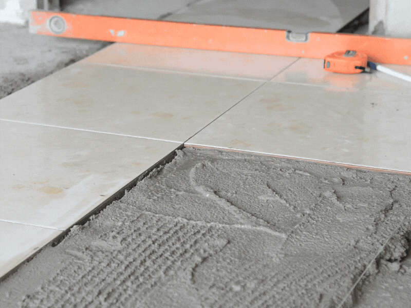 Close-up of white ceramic floor tiles being installed with gray adhesive mortar. An orange leveling tool and measuring tape are visible in the background, ensuring proper alignment. A trowel mark is visible in the mortar, indicating ongoing work to set the tiles evenly.