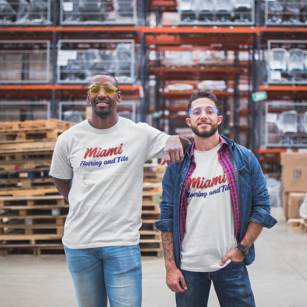 Two men in a warehouse, both wearing Miami Flooring and Tile t-shirts. The man on the left has yellow-tinted glasses and jeans, while the man on the right has a cap, checkered shirt over a white tee, and jeans. They stand in front of stacked pallets and shelves with boxed items.